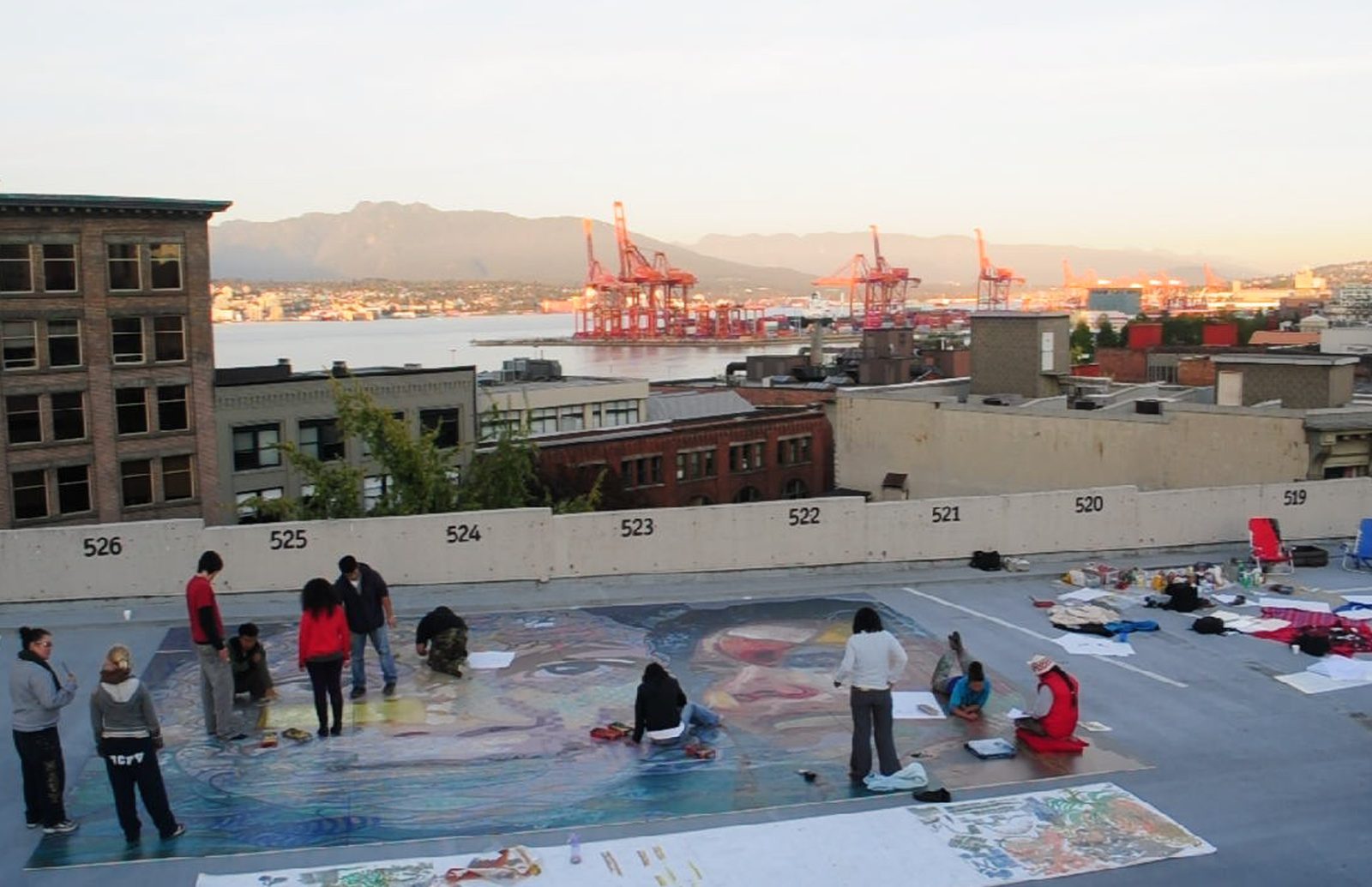 Participants painting The Belonging Action flat on a downtown Vancouver rooftop, the port cranes and the North Shore mountains behind them