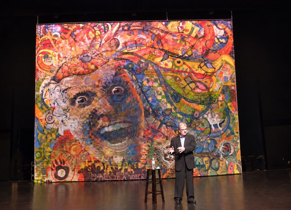 Sir Ken Robinson speaking on a theatre stage in front of the finished Out of Our Minds mural, which reads This is not a mural, this is a voice