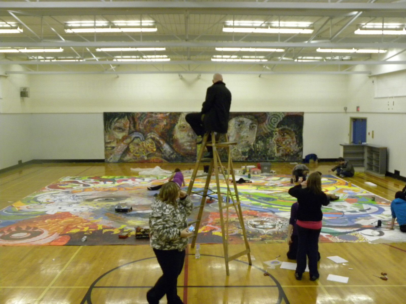 Scott Saywell on a stepladder painting the upper edge of Out of Our Minds, the rest of the painting laid out across the gymnasium floor around him